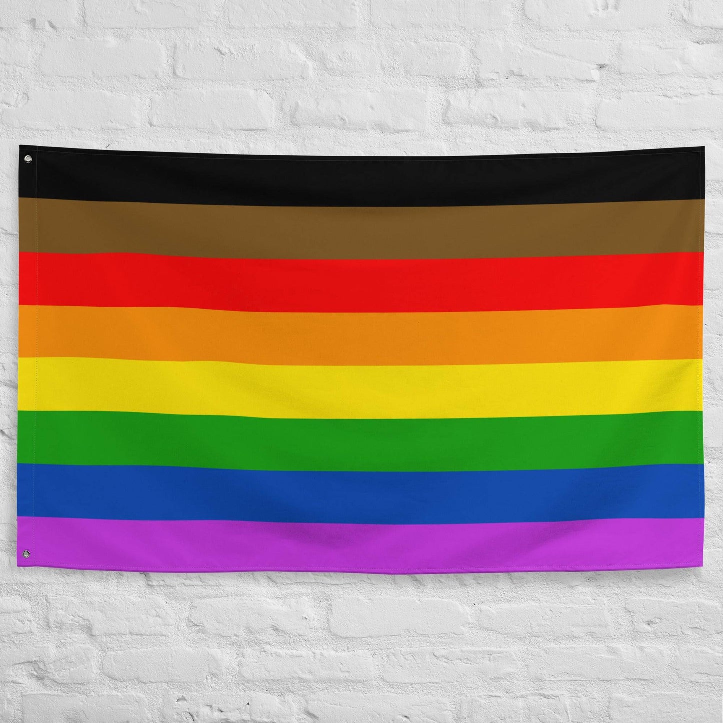 The Philadelphia Pride Flag by Pridelity, featuring horizontal stripes in black, brown, red, orange, yellow, green, blue, and purple, hangs against a white brick wall. This vibrant emblem of diversity and inclusion adds a splash of color reminiscent of a lively rainbow shop.