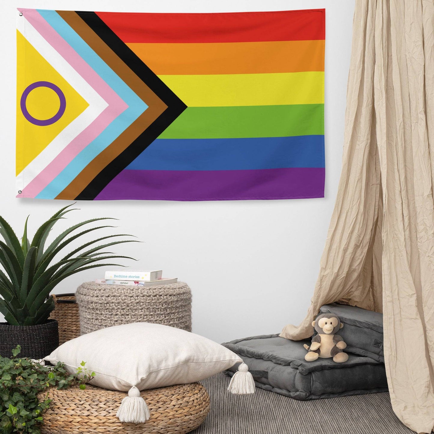 The image displays an Intersex-Inclusive Progress Pride Flag by Pridelity, hung on a white brick wall outside a vibrant rainbow shop. The flag features horizontal rainbow stripes and a chevron on the left, incorporating black, brown, light blue, pink, and white colors along with a purple circle on a yellow triangle.