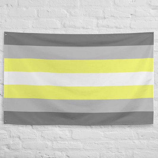 A Demigender Pride Flag from Pridelity, featuring horizontal stripes in gray, yellow, white, light yellow, and gray, adorns the textured white brick wall backdrop of a rainbow shop.