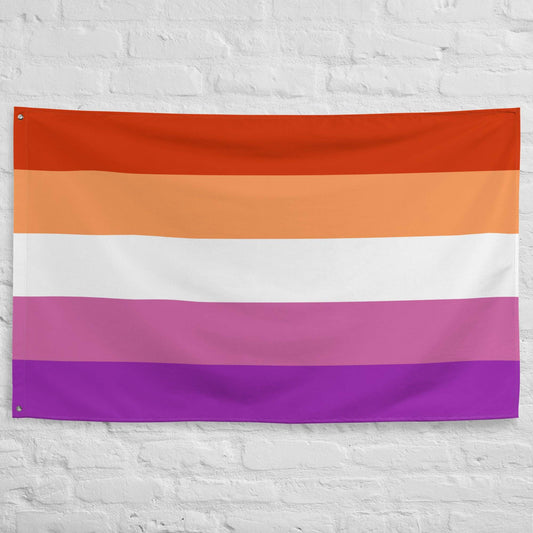 A Pridelity Lesbian Pride Flag with horizontal stripes in dark orange, orange, light orange, white, pink, and purple hung on a white brick wall outside the Rainbow Shop.