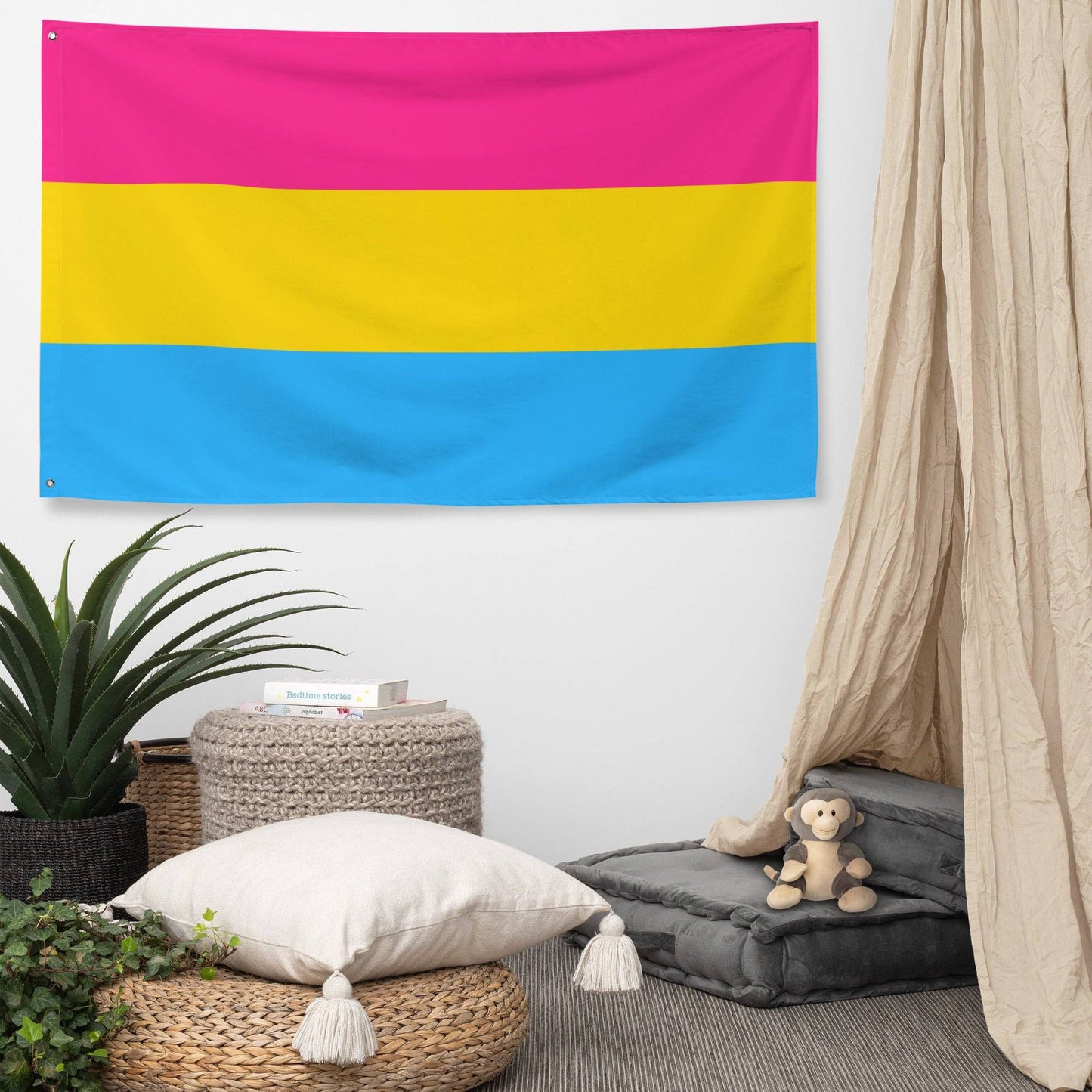 The Pan Pride Flag by Pridelity, featuring three horizontal stripes, hangs against a white brick wall. The top stripe is pink, the middle stripe is yellow, and the bottom stripe is blue, resembling a vibrant rainbow shop display.
