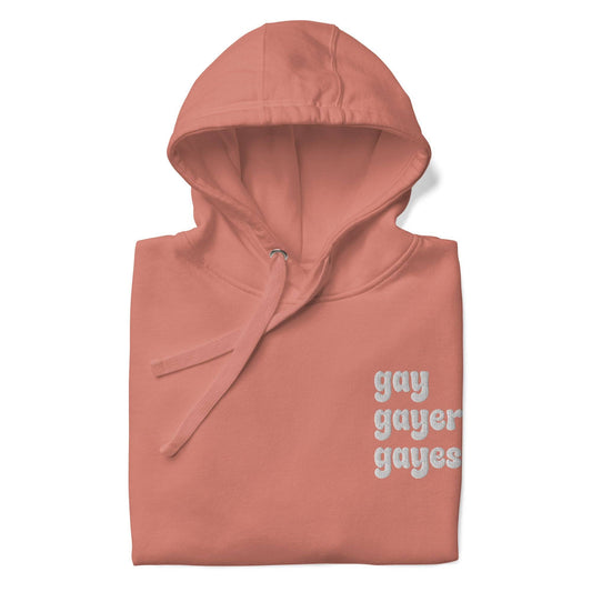 A Dusty Rose-colored hoodie from Pridelity's collection, named "Gay Gayer Gayest Hoodie," features a front pocket and the text "gay gayer gayest" printed in white on the upper left side.
