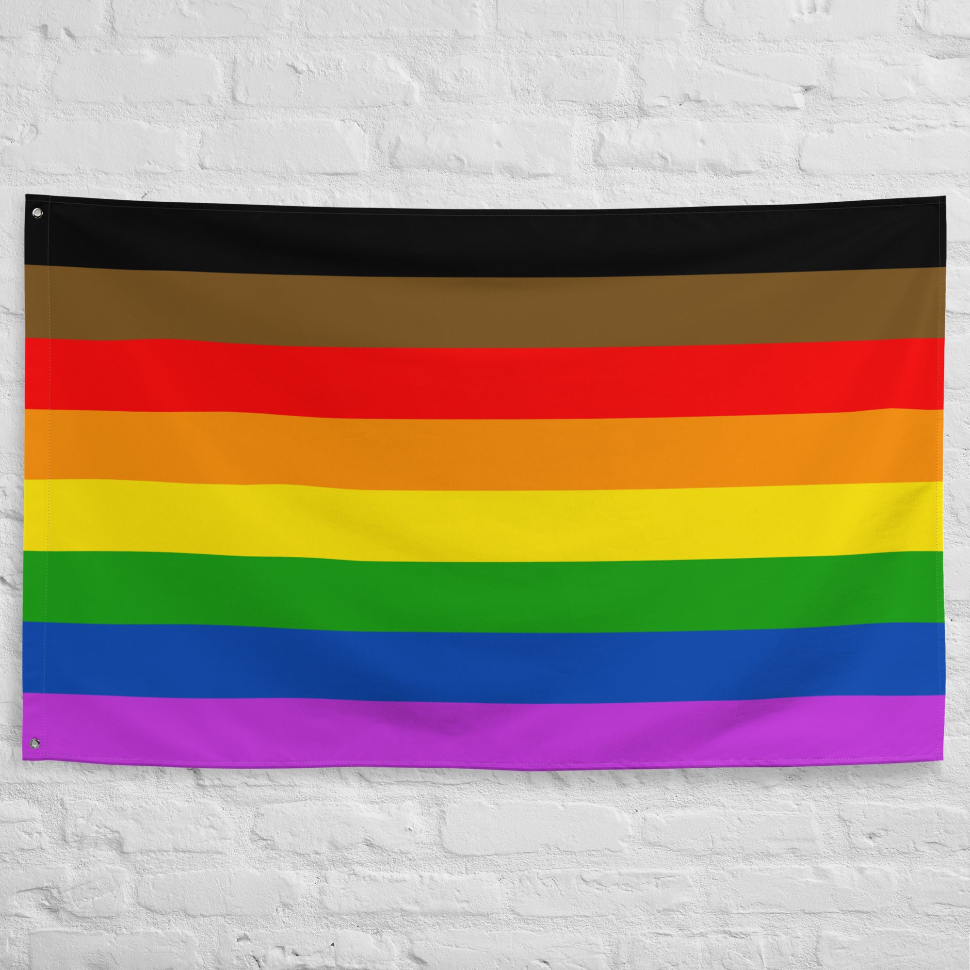 The Philadelphia Pride Flag by Pridelity, featuring horizontal stripes in black, brown, red, orange, yellow, green, blue, and purple, hangs against a white brick wall. This vibrant emblem of diversity and inclusion adds a splash of color reminiscent of a lively rainbow shop.