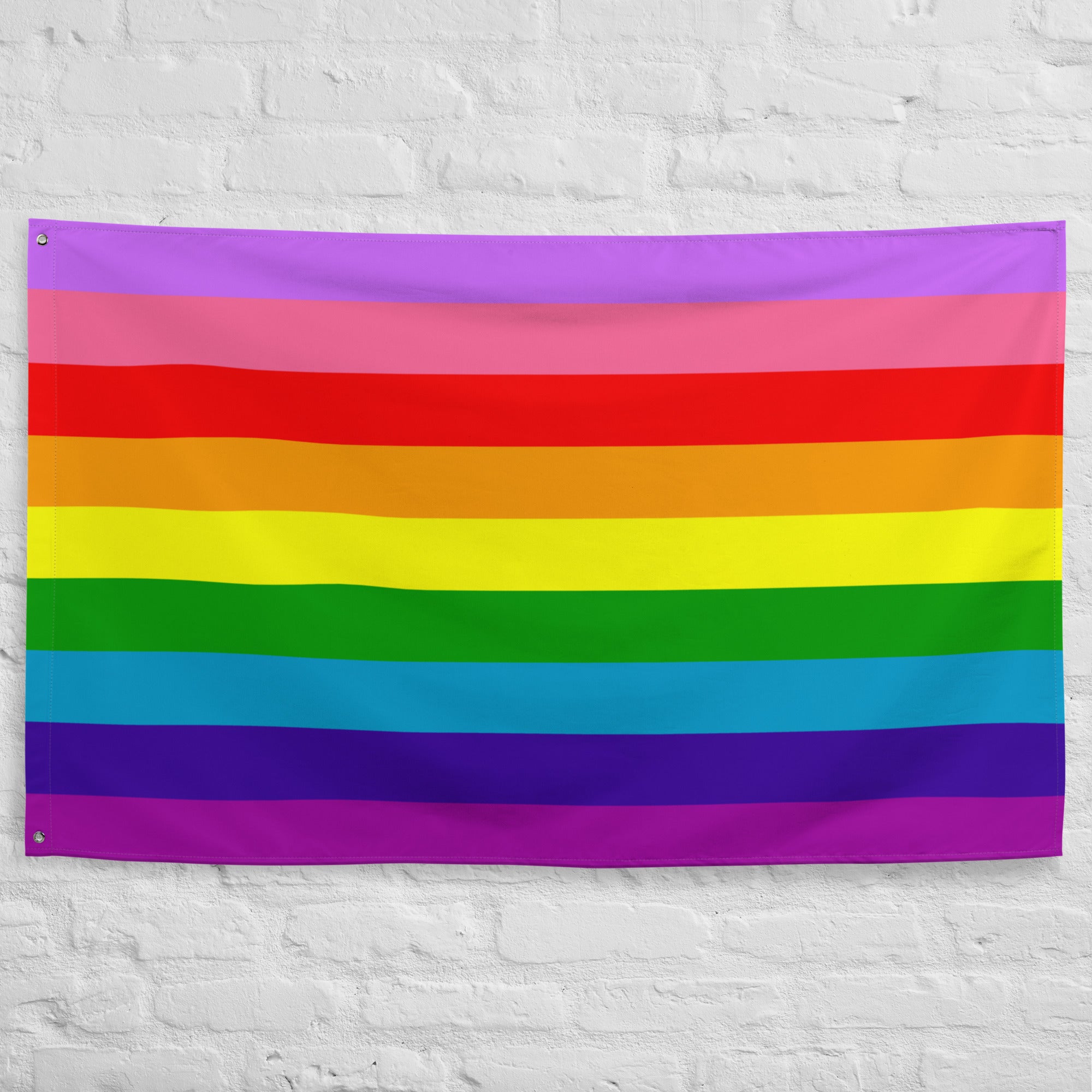 The Gilbert Baker Pride Flag from Pridelity, featuring vibrant horizontal stripes in red, orange, yellow, green, blue, and purple, elegantly adorns a white brick wall.