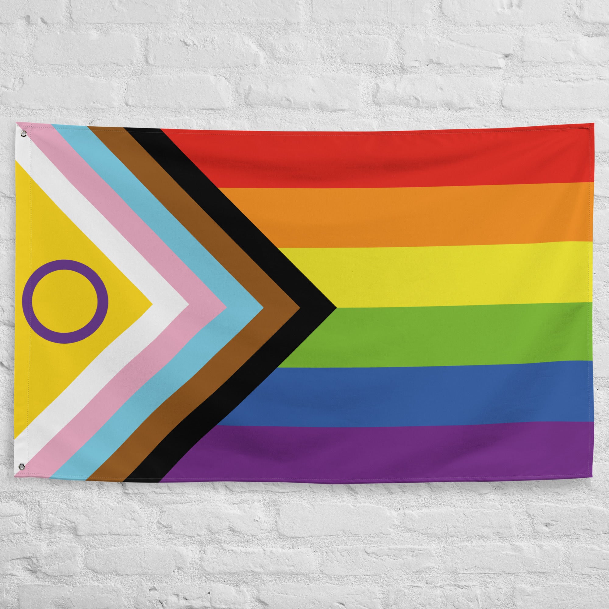 The image displays an Intersex-Inclusive Progress Pride Flag by Pridelity, hung on a white brick wall outside a vibrant rainbow shop. The flag features horizontal rainbow stripes and a chevron on the left, incorporating black, brown, light blue, pink, and white colors along with a purple circle on a yellow triangle.