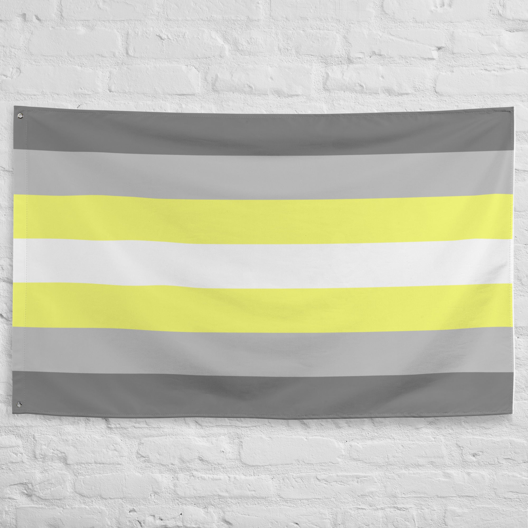 A Demigender Pride Flag from Pridelity, featuring horizontal stripes in gray, yellow, white, light yellow, and gray, adorns the textured white brick wall backdrop of a rainbow shop.