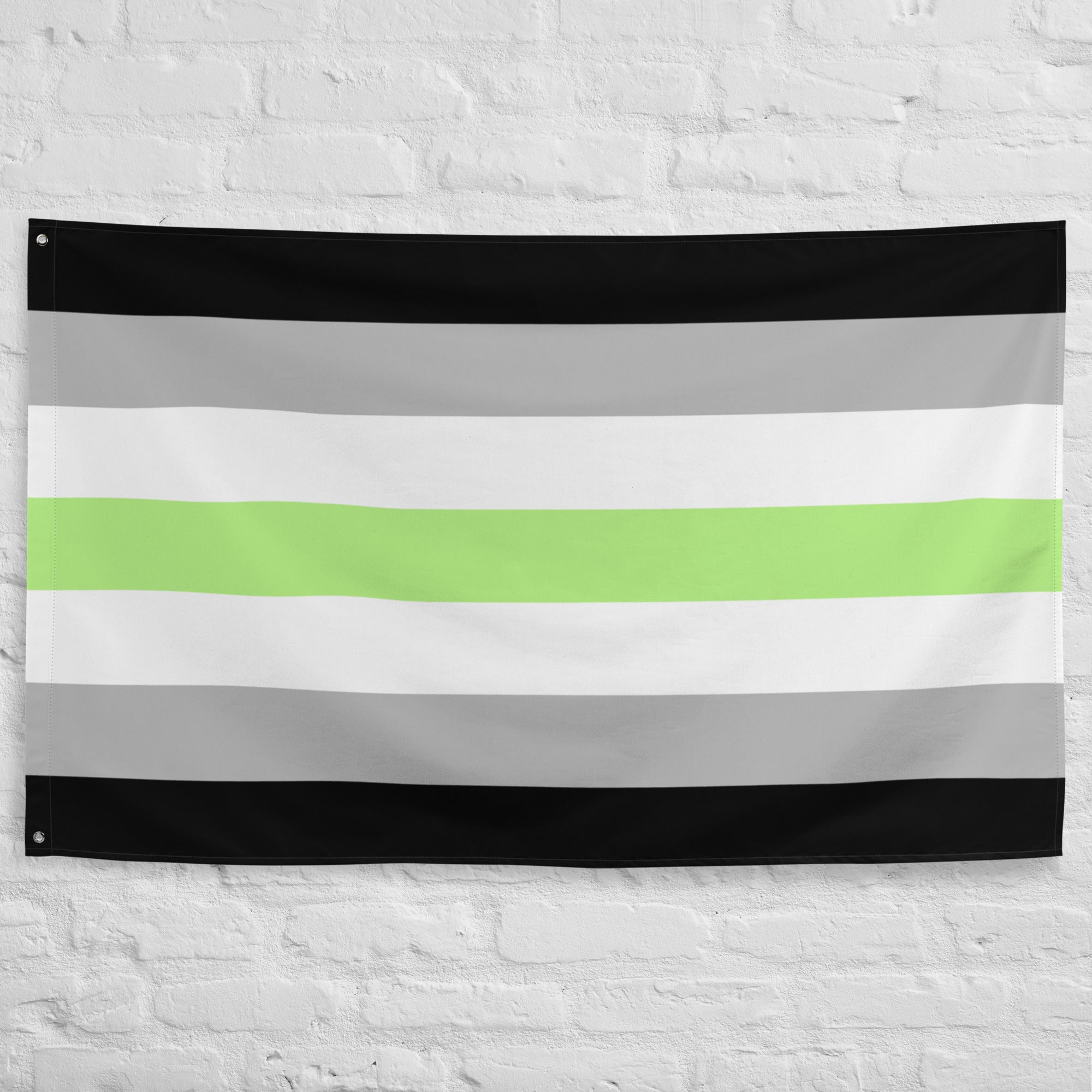 The Agender Pride Flag by Pridelity, showcasing horizontal stripes in black, gray, white, a central green stripe, followed by white, gray, and black once more, is prominently displayed as part of our Pride Collections against a white brick wall.