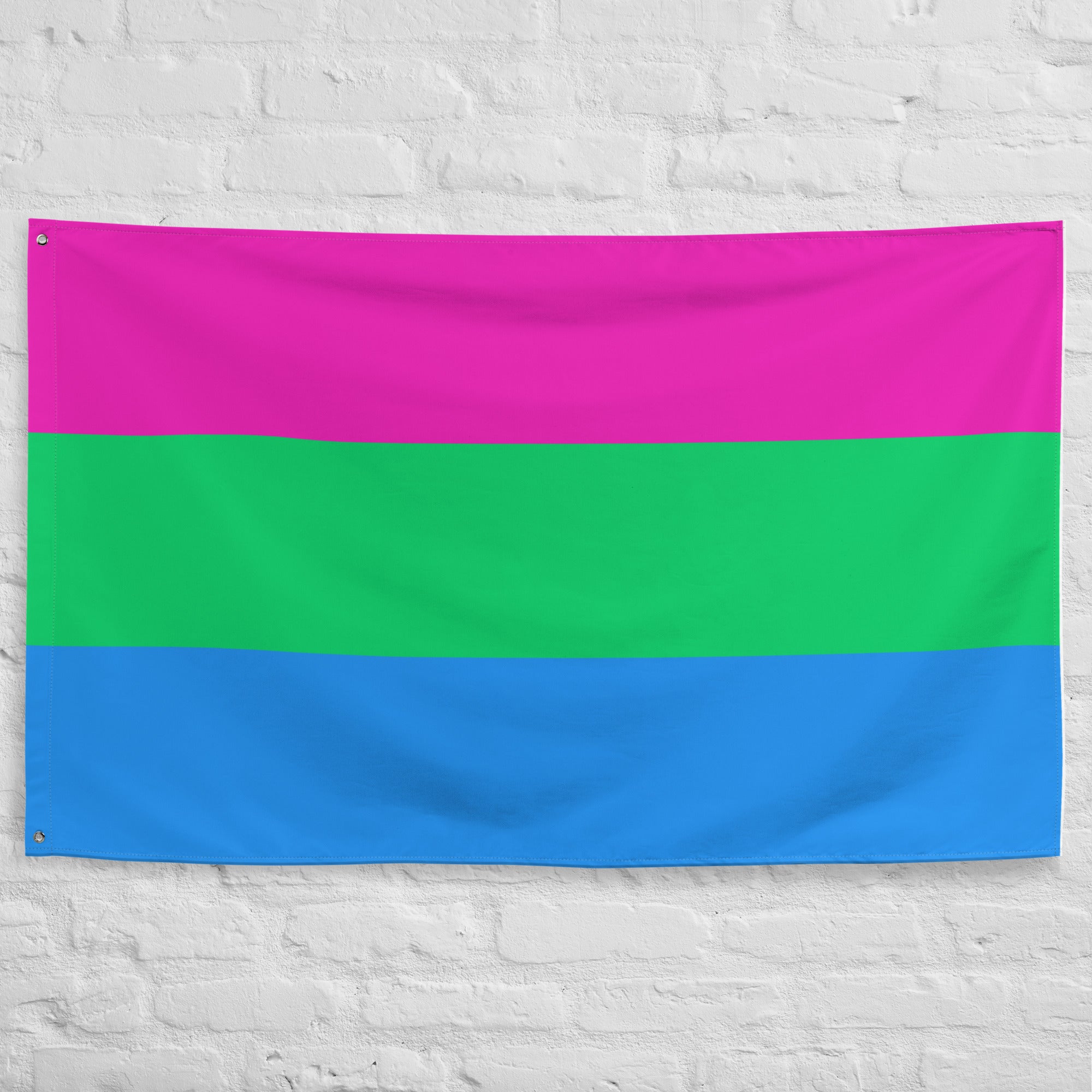 A vibrant Polysexual Flag, featuring three horizontal stripes with pink on top, green in the middle, and blue at the bottom, hangs elegantly on a white brick wall. From Pridelity, it adds a splash of color reminiscent of a rainbow shop display.