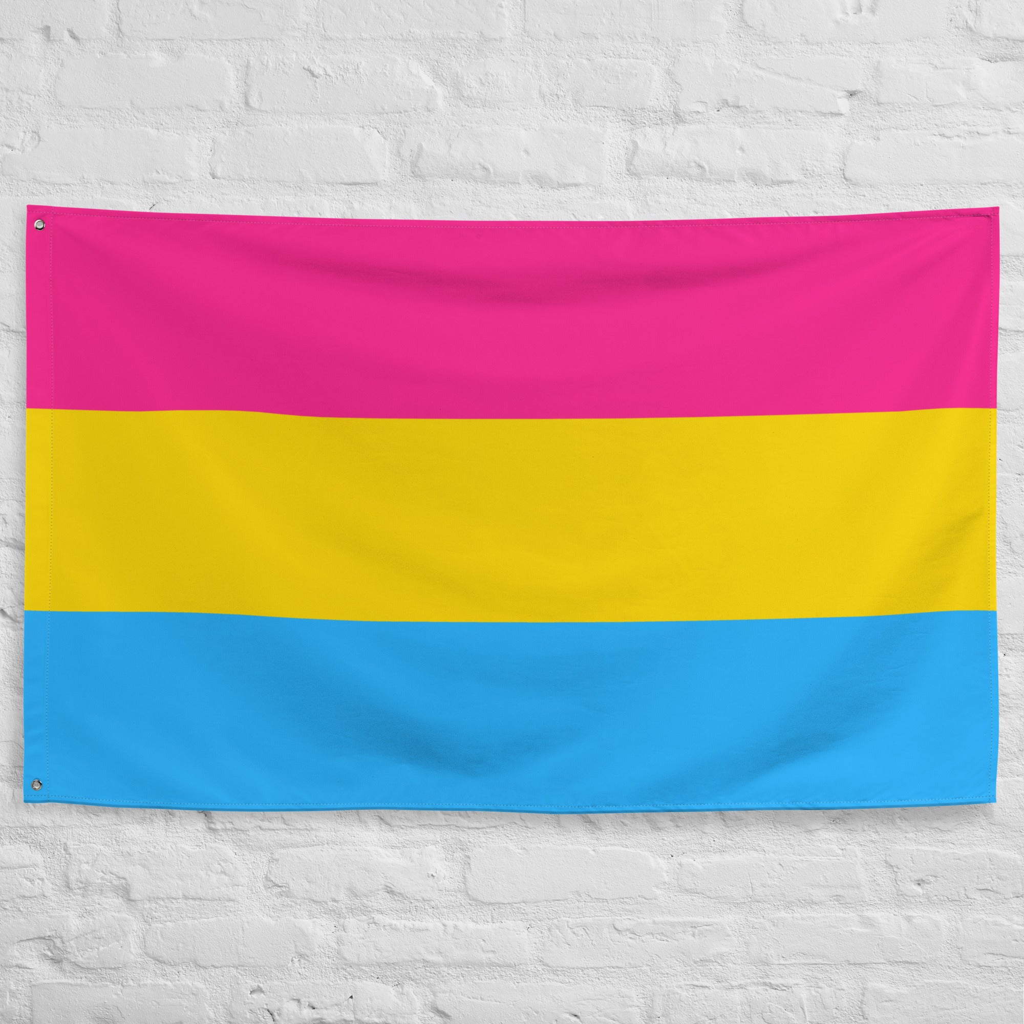 The Pan Pride Flag by Pridelity, featuring three horizontal stripes, hangs against a white brick wall. The top stripe is pink, the middle stripe is yellow, and the bottom stripe is blue, resembling a vibrant rainbow shop display.