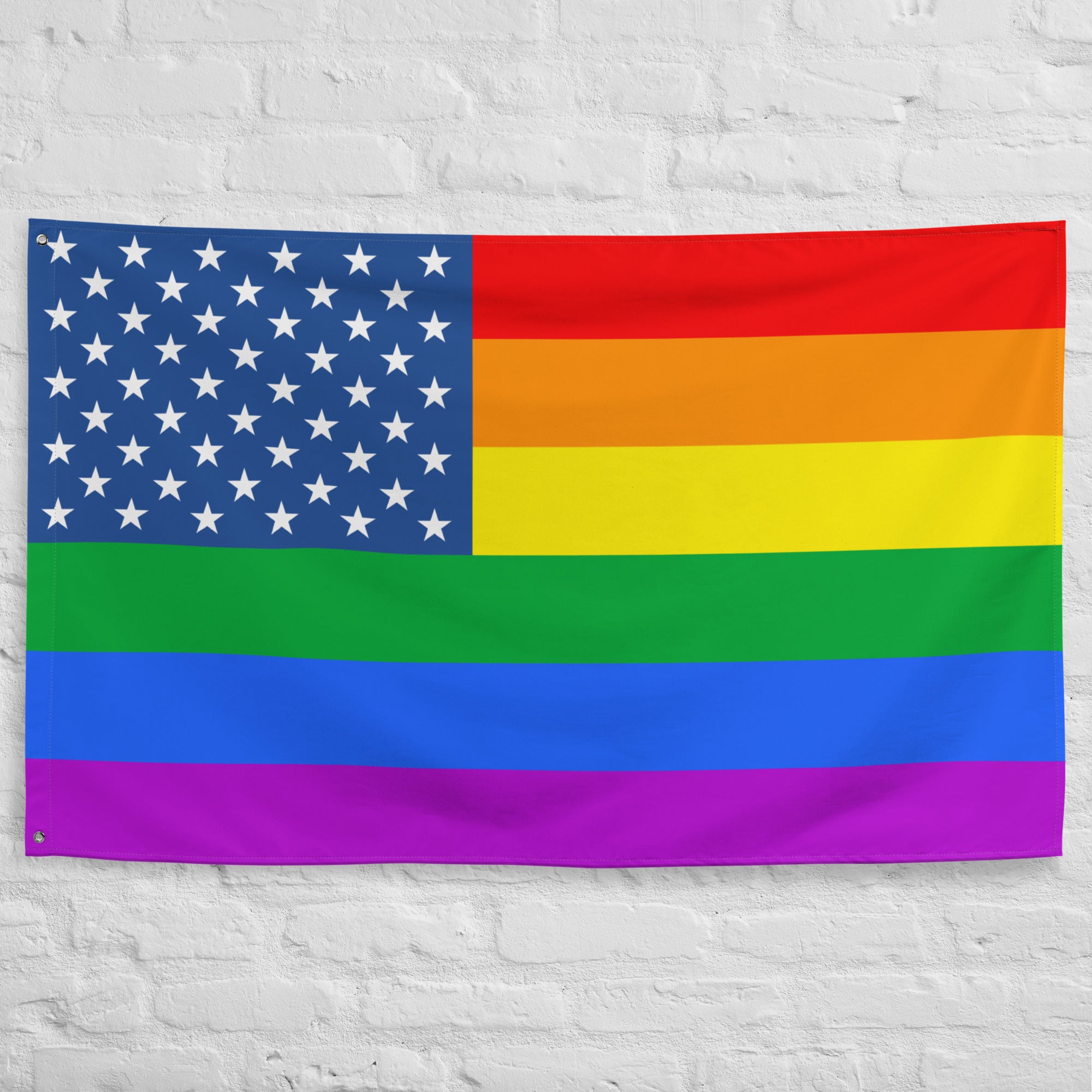 The Pride Stars and Stripes Flag from Pridelity, featuring an American flag seamlessly merged with the vibrant rainbow stripes of the LGBTQ+ pride flag, hangs against a white brick wall. Its stars on a blue background in the top left beautifully contrast with the horizontal rainbow stripes, creating an inviting and colorful display.